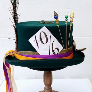 May include: A green top hat with a gold band and a white card with the number "10/6" written on it. The hat has a peacock feather and gold and colorful beads on top. The hat is sitting on a wooden stand with a purple and orange ribbon.
