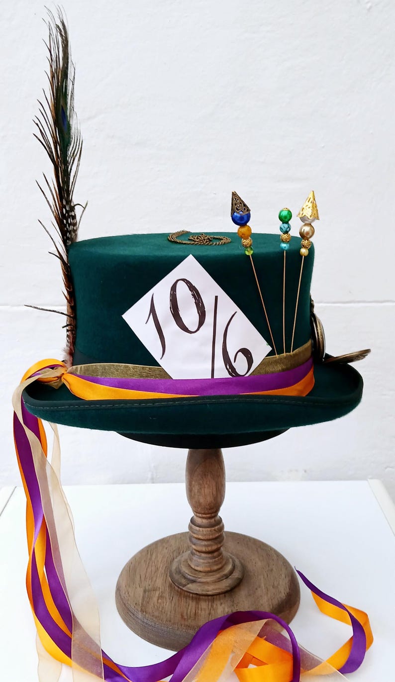 May include: A green top hat with a gold band and a white card with the number "10/6" written on it. The hat has a peacock feather and gold and colorful beads on top. The hat is sitting on a wooden stand with a purple and orange ribbon.