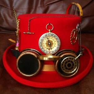 Puede incluir: Un sombrero de copa steampunk rojo con una br&uacute;jula, gafas y accesorios decorativos. El sombrero presenta una cinta dorada, un peque&ntilde;o reloj de arena y una cadena. Las gafas tienen lentes oscuras y son de metal color cobre.