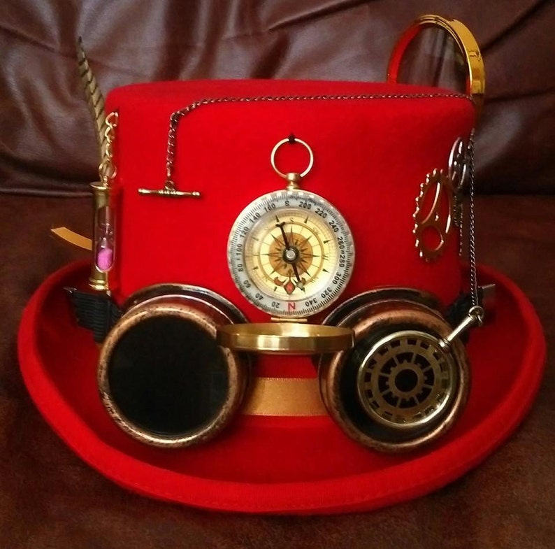 Puede incluir: Un sombrero de copa steampunk rojo con una br&uacute;jula, gafas y accesorios decorativos. El sombrero presenta una cinta dorada, un peque&ntilde;o reloj de arena y una cadena. Las gafas tienen lentes oscuras y son de metal color cobre.