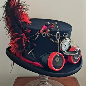 May include: A black top hat with a red feather, a chain with beads, a pocket watch, and goggles with red lenses. The hat is on a wooden stand.