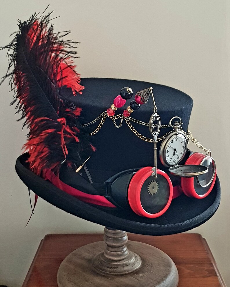 May include: A black top hat with a red feather, a chain with beads, a pocket watch, and goggles with red lenses. The hat is on a wooden stand.