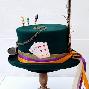 May include: A green top hat with a gold chain, playing cards, and a feather. The hat is on a wooden stand with orange and purple ribbons.