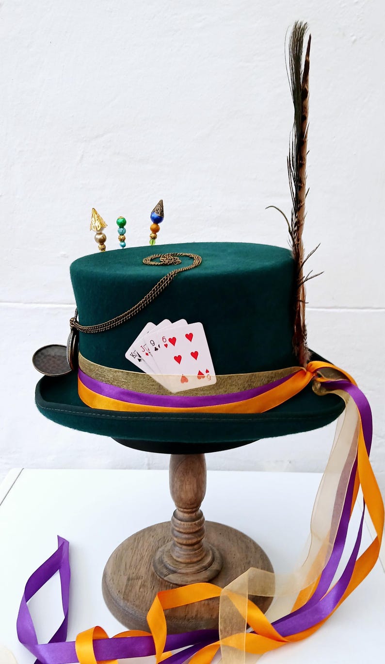 May include: A green top hat with a gold chain, playing cards, and a feather. The hat is on a wooden stand with orange and purple ribbons.