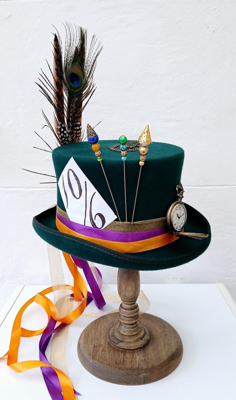May include: A green top hat with a purple and orange ribbon, a pocket watch, and three pins with beads. The hat has a white card attached to the side that says "10/6". The hat is on a wooden stand with a brown base and a round top. There are orange and purple ribbons hanging from the hat.