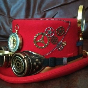 Puede incluir: Un sombrero steampunk rojo con engranajes de lat&oacute;n, una br&uacute;jula y gafas. Una lupa est&aacute; adjunta. El sombrero tiene una banda el&aacute;stica negra y una cinta dorada.