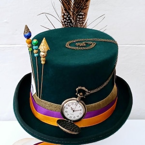 May include: A green top hat with a purple and orange ribbon, a gold chain with a pocket watch, and a peacock feather on top. The hat is on a wooden stand.