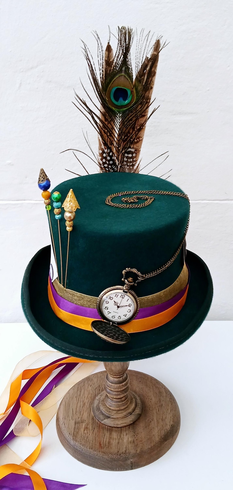 May include: A green top hat with a purple and orange ribbon, a gold chain with a pocket watch, and a peacock feather on top. The hat is on a wooden stand.