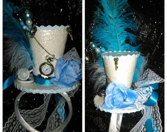 May include: A whimsical, miniature top hat in white, adorned with blue feathers, a blue paper rose, and a vintage pocket watch. The hat features a key charm, lace trim, and a silver-colored neck ring.