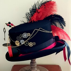 May include: A black top hat with a red and black feather plume. The hat is decorated with gold gears and a key. The hat is sitting on a wooden stand.