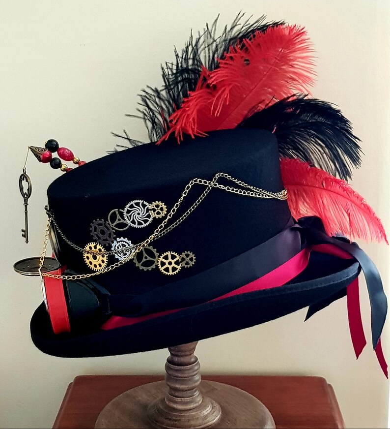 May include: A black top hat with a red and black feather plume. The hat is decorated with gold gears and a key. The hat is sitting on a wooden stand.