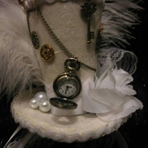 Puede incluir: Un sombrero de copa blanco con una pluma blanca, una flor blanca, un engranaje dorado, una llave y un reloj de bolsillo con una cadena.