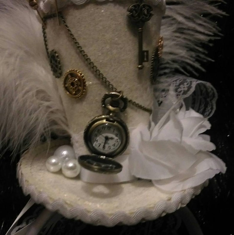 Puede incluir: Un sombrero de copa blanco con una pluma blanca, una flor blanca, un engranaje dorado, una llave y un reloj de bolsillo con una cadena.