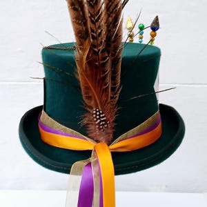 May include: A green top hat with a gold band and a purple and orange ribbon. The hat is decorated with brown feathers and colorful beads.