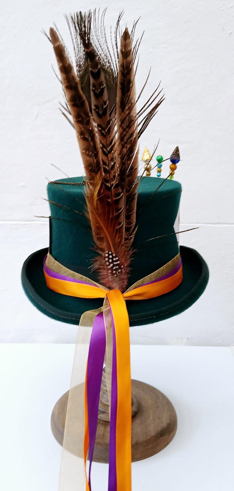 May include: A green top hat with a gold band and a purple and orange ribbon. The hat is decorated with brown feathers and colorful beads.