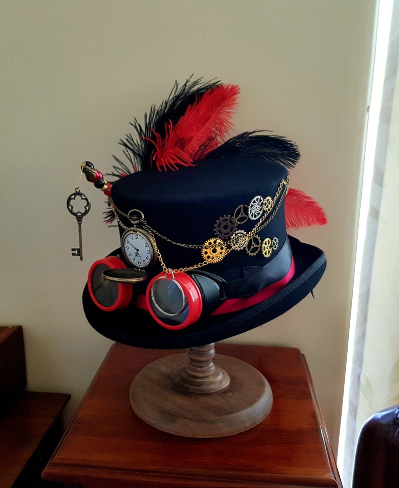 May include: A black top hat with a red and black feather, gold chain, and gears. The hat also has a pocket watch and goggles with red lenses.