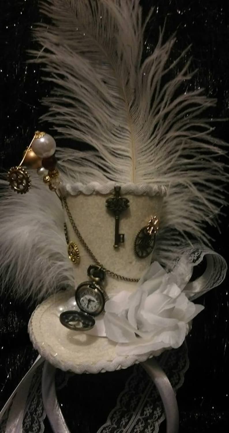 Puede incluir: Un sombrero de copa blanco con una pluma blanca, un dije de llave, un reloj de bolsillo y una rosa blanca. El sombrero est&aacute; decorado con engranajes y cadenas doradas.