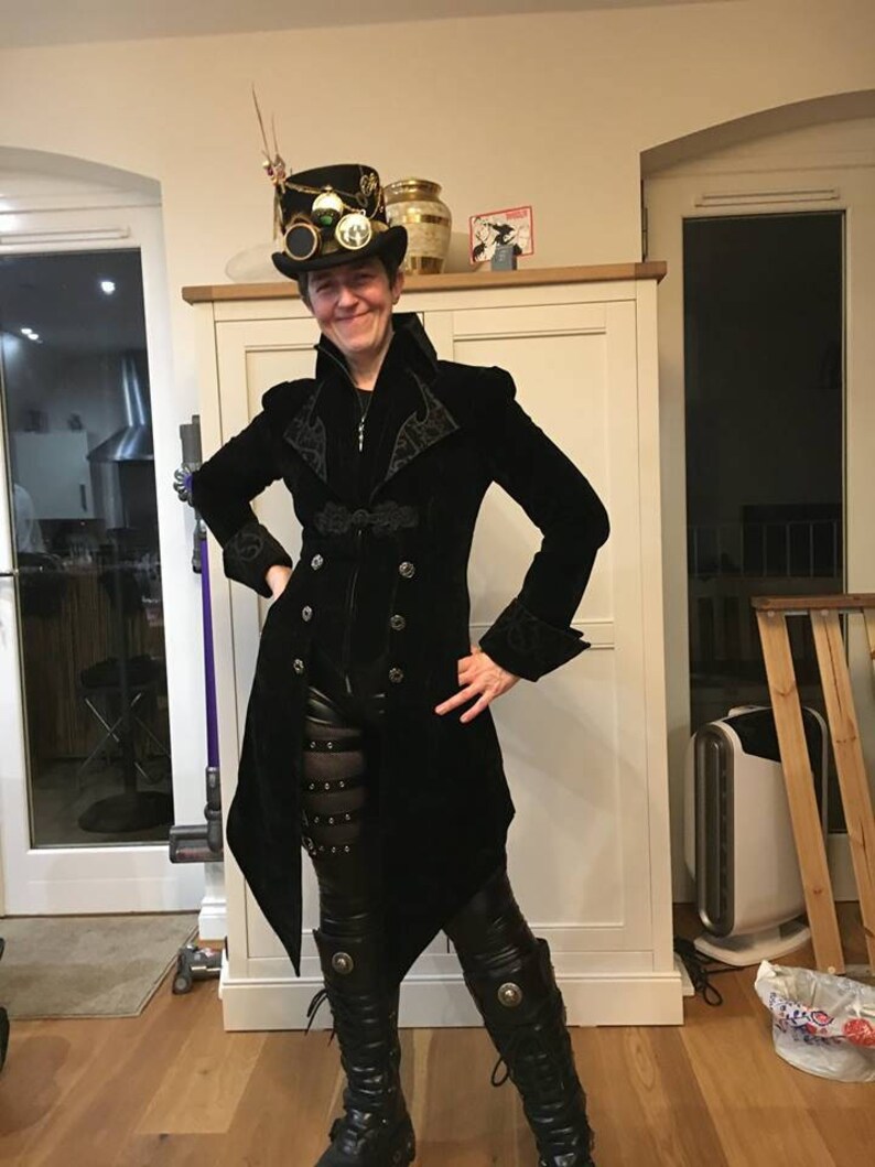 May include: A person wearing a black velvet coat with gold buttons and a black top hat with steampunk-style embellishments. The person is wearing black leather pants and black boots.