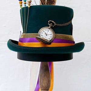 May include: A green top hat with a gold pocket watch, purple and orange ribbon, and a peacock feather. The hat is on a wooden stand.