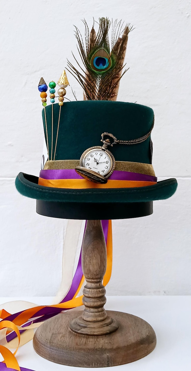 May include: A green top hat with a gold pocket watch, purple and orange ribbon, and a peacock feather. The hat is on a wooden stand.
