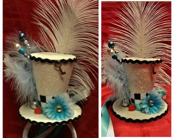 May include: A whimsical mini top hat with a white sequined body, black scalloped trim, and a black and white checkered band. Adorned with a blue flower, feathers, and decorative beads. A key charm hangs from the hat.