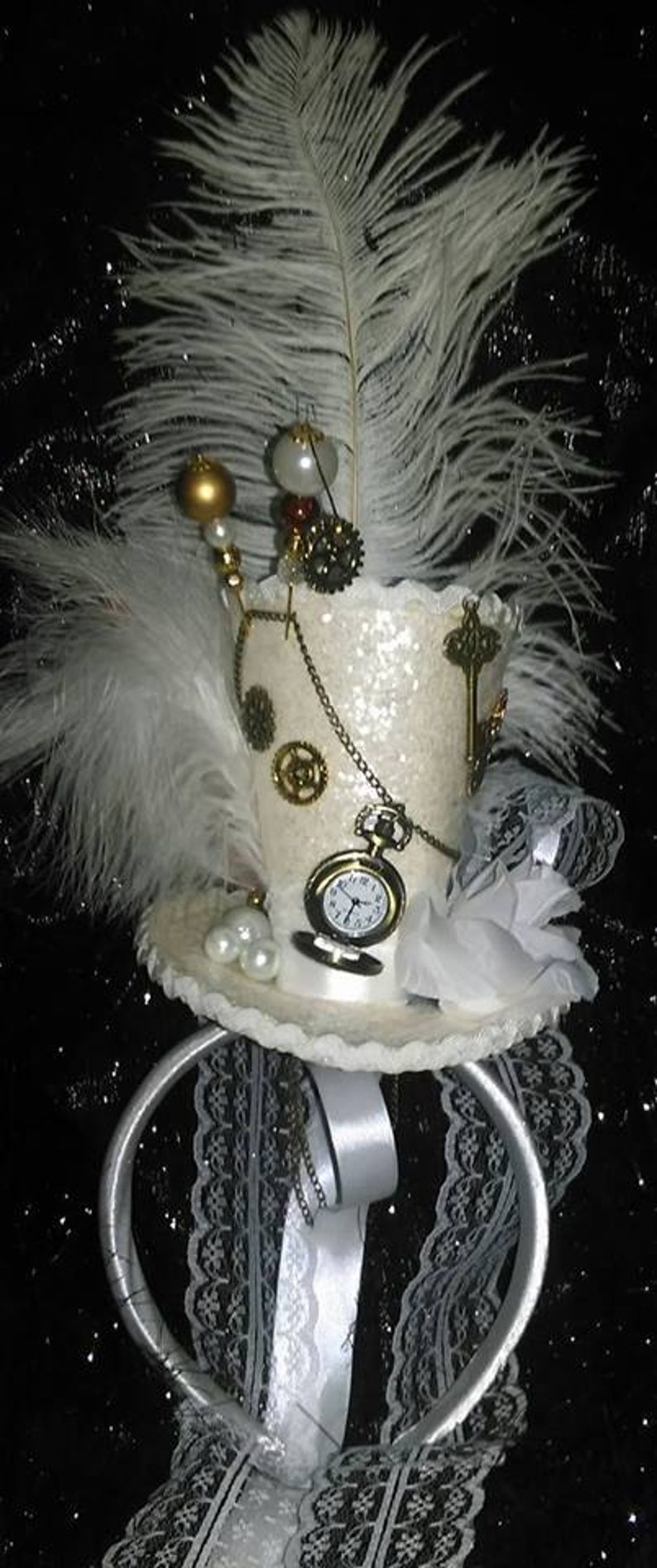 Puede incluir: Sombrero de copa blanco con una pluma blanca, un reloj de bolsillo y una cadena. El sombrero est&aacute; decorado con purpurina, perlas y un ribete de encaje. El sombrero est&aacute; sobre un fondo negro.