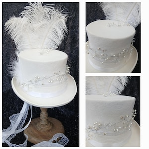 Puede incluir: Un sombrero de copa blanco con ala ancha, adornado con plumas blancas, una vid plateada con cuentas y una cinta blanca. El sombrero se exhibe sobre un soporte de madera, mostrando su elegante diseño. Perfecto para bodas o eventos especiales.