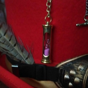 Puede incluir: Un sombrero de copa de fieltro rojo con un colgante de reloj de arena dorado, una pluma y gafas de estilo steampunk. El reloj de arena contiene arena rosa. El sombrero tiene una banda negra y un ribete dorado.