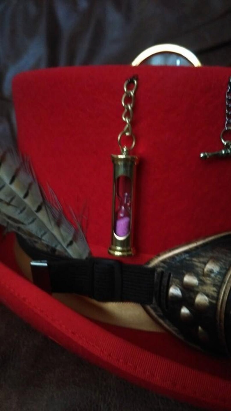 Puede incluir: Un sombrero de copa de fieltro rojo con un colgante de reloj de arena dorado, una pluma y gafas de estilo steampunk. El reloj de arena contiene arena rosa. El sombrero tiene una banda negra y un ribete dorado.