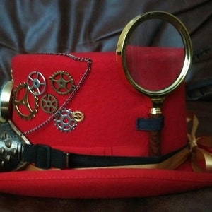 Puede incluir: Un sombrero de copa steampunk rojo con engranajes de lat&oacute;n, una lupa y gafas. El sombrero presenta una correa negra, una cinta dorada y una cadena. Los engranajes son de varios tama&ntilde;os y colores.
