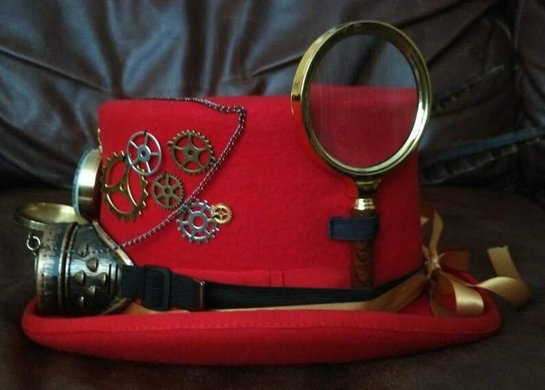 Puede incluir: Un sombrero de copa steampunk rojo con engranajes de lat&oacute;n, una lupa y gafas. El sombrero presenta una correa negra, una cinta dorada y una cadena. Los engranajes son de varios tama&ntilde;os y colores.