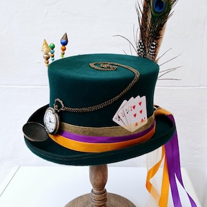May include: A green top hat with a gold chain, pocket watch, playing cards, and a peacock feather. The hat is on a wooden stand with orange and purple ribbons.