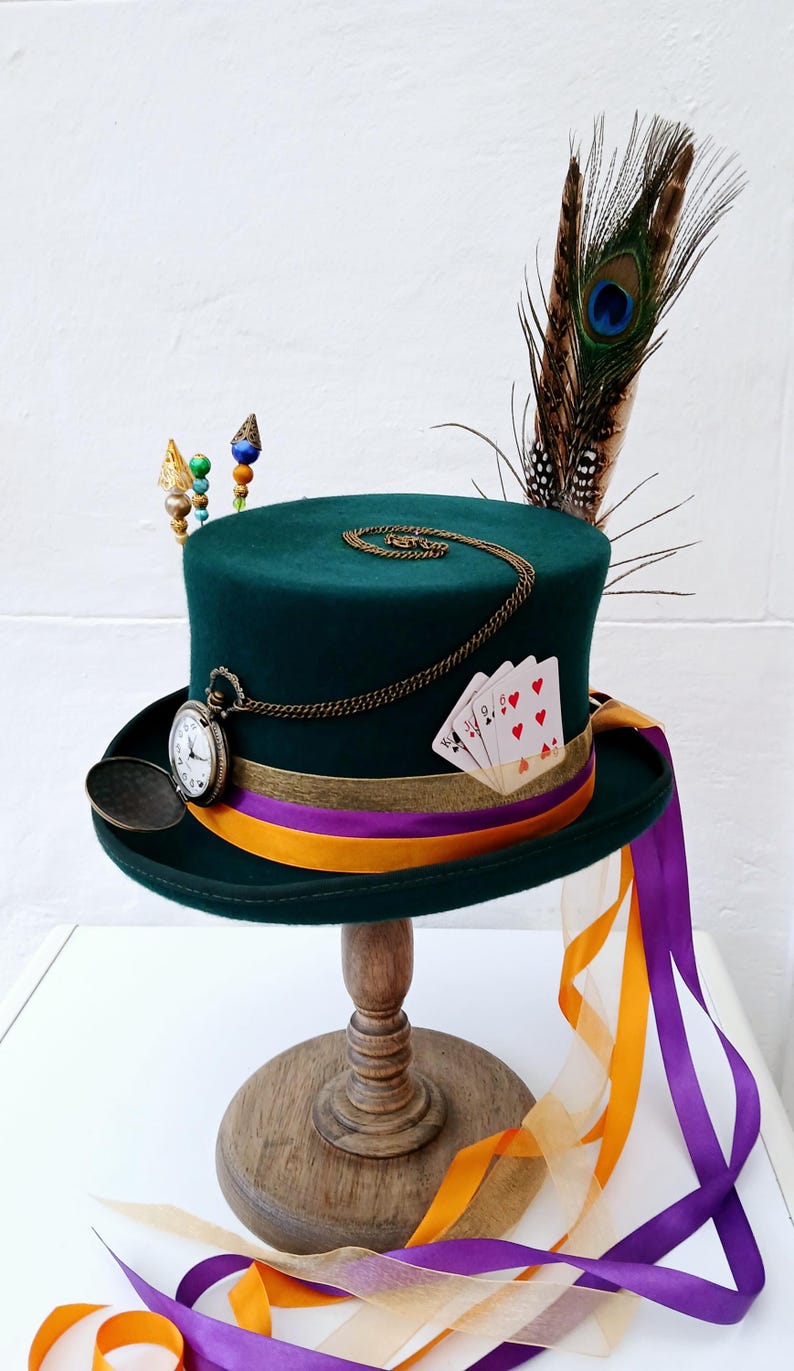 May include: A green top hat with a gold chain, pocket watch, playing cards, and a peacock feather. The hat is on a wooden stand with orange and purple ribbons.