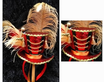 Peut inclure: Un mini chapeau haut-de-forme rouge, noir et doré, orné de paillettes, de chaînes dorées et d'une grande plume. Le chapeau a un petit bord et est fixé à un bandeau avec des rubans rouges et dorés.