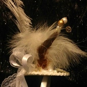 Puede incluir: Una diadema de plumas blancas con un borde de encaje blanco y un adorno blanco y dorado. La diadema est&aacute; decorada con perlas y un peque&ntilde;o engranaje.