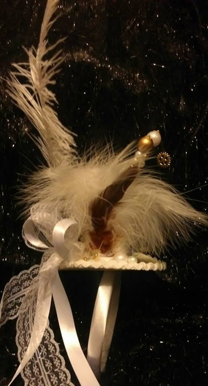 Puede incluir: Una diadema de plumas blancas con un borde de encaje blanco y un adorno blanco y dorado. La diadema est&aacute; decorada con perlas y un peque&ntilde;o engranaje.