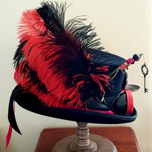 May include: A black top hat with a red and black feather, a gold chain, and a key charm. The hat is decorated with red and black beads and has a steampunk-style design.