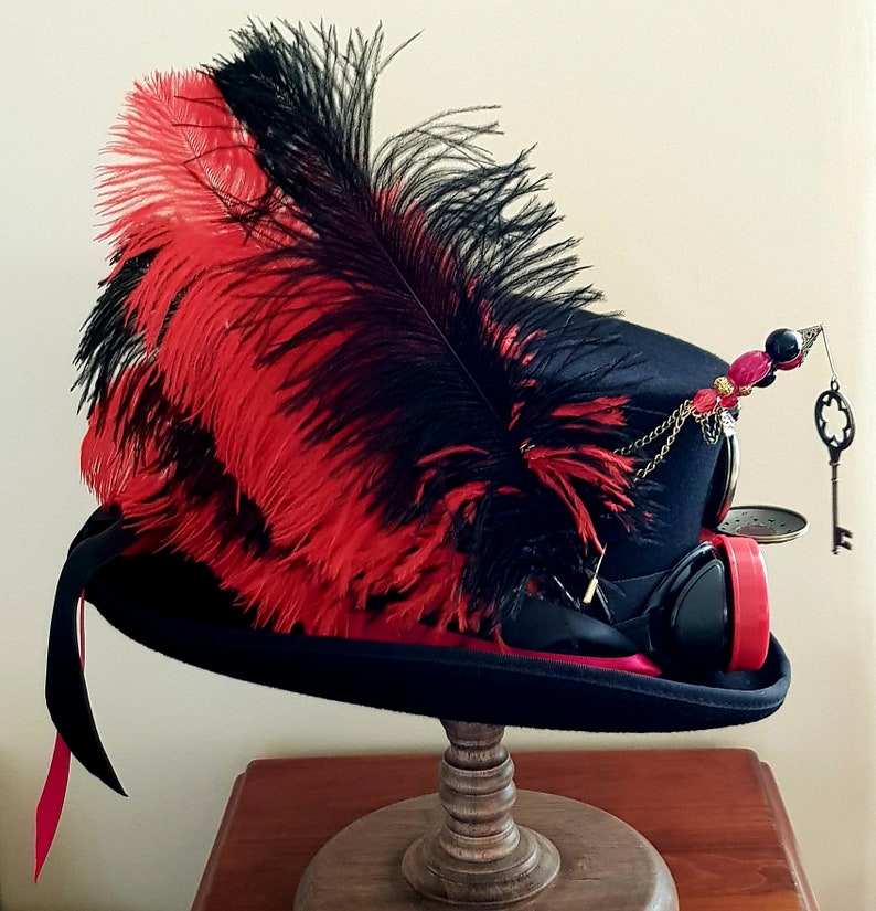 May include: A black top hat with a red and black feather, a gold chain, and a key charm. The hat is decorated with red and black beads and has a steampunk-style design.