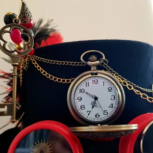May include: A black top hat with a gold chain, a key, and a pocket watch with a white face and black hands. The watch face has black Roman numeral markings and the words "QUARTZ" and "JAPAN" printed on it.