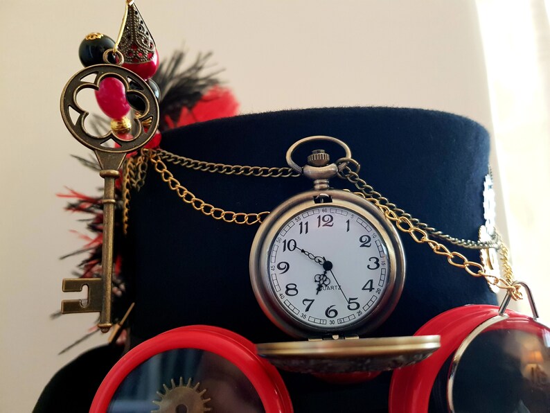 May include: A black top hat with a gold chain, a key, and a pocket watch with a white face and black hands. The watch face has black Roman numeral markings and the words "QUARTZ" and "JAPAN" printed on it.