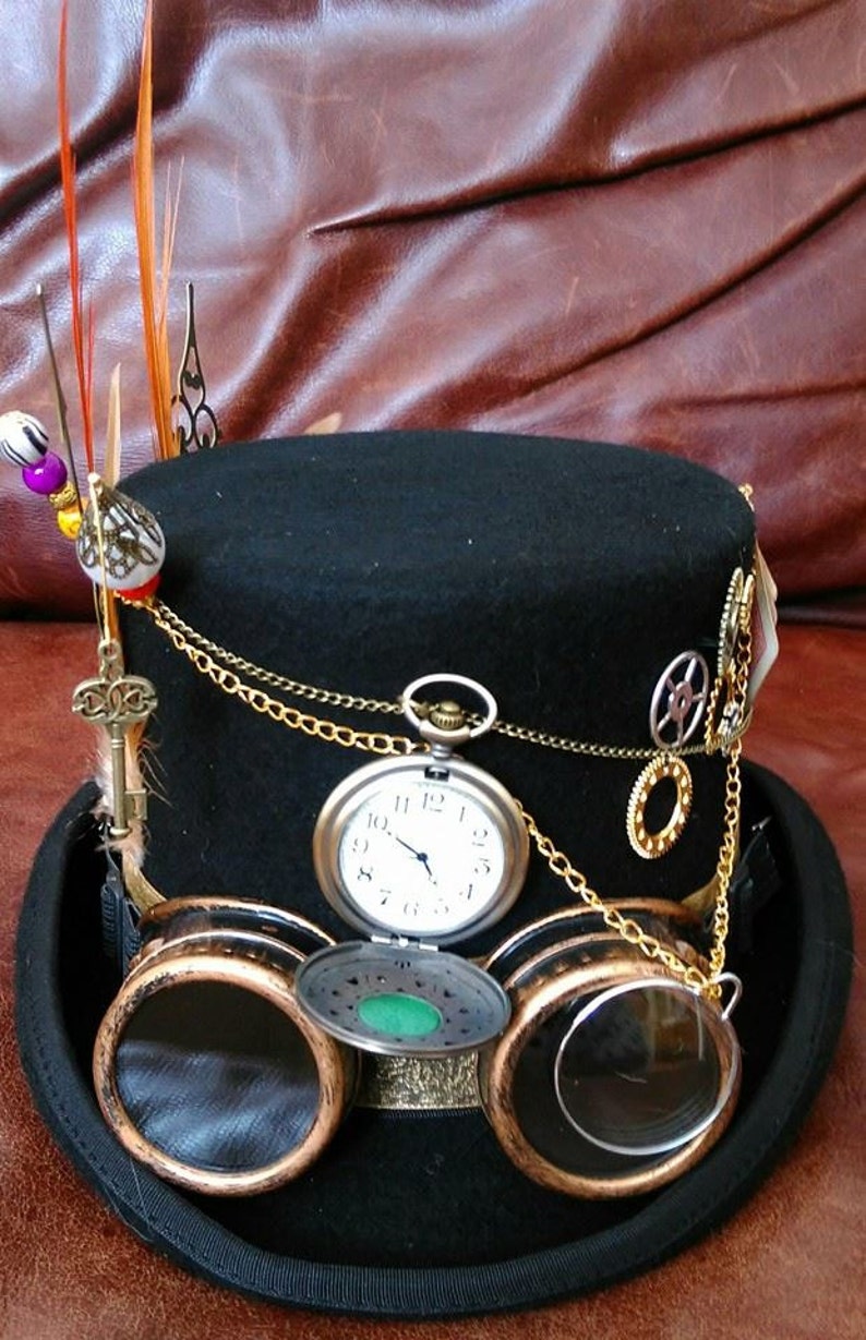 May include: A black top hat with gold chains, a pocket watch, and goggles. The hat has a feather on the side and a small key charm.