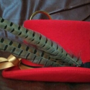 Puede incluir: Un sombrero rojo con una cinta dorada, adornado con plumas moteadas. Una peque&ntilde;a cadena dorada con un dije de reloj de arena en miniatura y un dije redondo cuelgan del sombrero. El sombrero tiene un anillo dorado en la parte superior.