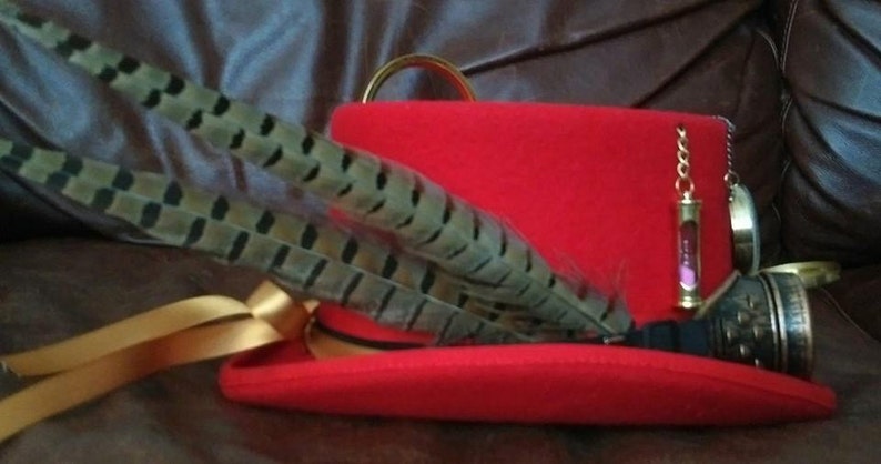 Puede incluir: Un sombrero rojo con una cinta dorada, adornado con plumas moteadas. Una peque&ntilde;a cadena dorada con un dije de reloj de arena en miniatura y un dije redondo cuelgan del sombrero. El sombrero tiene un anillo dorado en la parte superior.