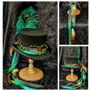 May include: A black top hat with a teal ribbon and gold embellishments. The hat features a peacock feather, brown and teal feathers, and a gold decorative element. The hat is displayed on a wooden stand.