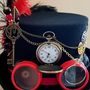 May include: A black top hat with a red and black feather, a gold chain, a pocket watch, and red goggles with a brass gear detail.
