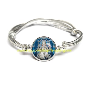 Puede incluir: Una pulsera de plata con una imagen redonda de la deidad hindú Ganesha, en azul y blanco. La imagen está enmarcada en plata y la pulsera está hecha de un cordón trenzado.
