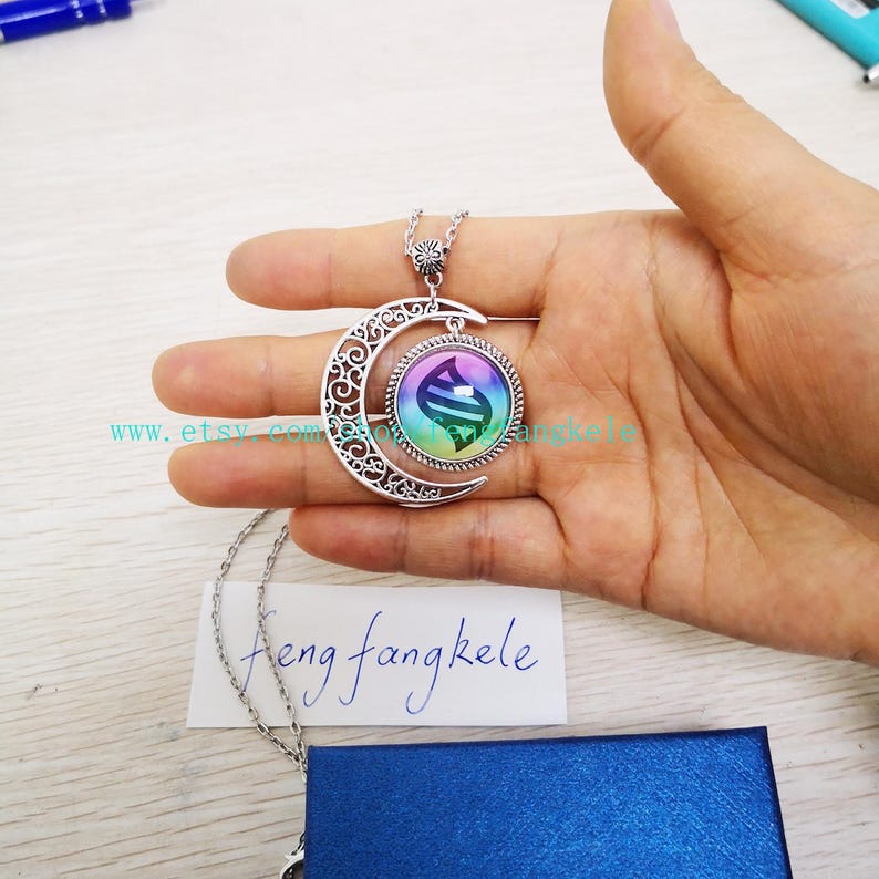 Op de afbeelding: Een zilveren halve maan ketting met een glazen hanger met een regenboogkleurige gradi&euml;nt en een zwart-wit symbool. De hanger is bevestigd aan een zilveren ketting.