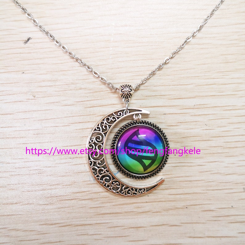 Op de afbeelding: Een zilveren halve maan ketting met een regenboogkleurige glazen hanger met een zwart-wit symbool. De hanger is bevestigd aan de halve maan met een zilveren ring.