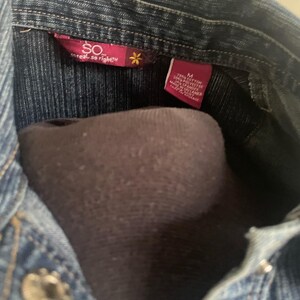 May include: A blue denim shirt with a pink label that reads "SO real, so right" and a small flower. The label also includes the size "M" and the fabric content: 70% cotton, 20% polyester, and 10% spandex. The shirt is made in Sri Lanka.