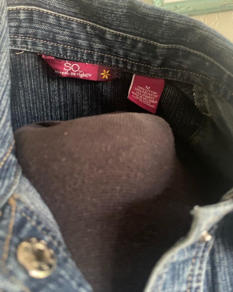 May include: A blue denim shirt with a pink label that reads "SO real, so right" and a small flower. The label also includes the size "M" and the fabric content: 70% cotton, 20% polyester, and 10% spandex. The shirt is made in Sri Lanka.